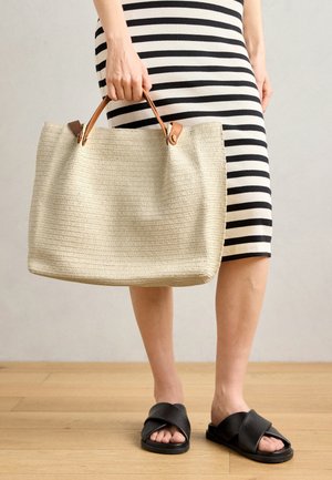 Beige vævet tote taske med brune læderhåndtag, der har en struktureret overflade. Kombineret med en sort og hvid stribet nederdel og sorte slidesandaler.