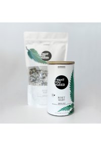 Contenitore cilindrico bianco con coperchio in legno, decorato con grafiche di foglie verdi e testo nero; una busta bianca con testo stampato e contenuti multicolori dietro di esso.