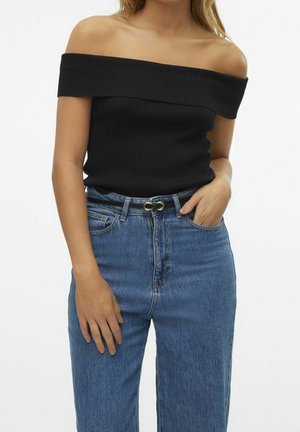 Vrouw draagt een zwarte geribde off-shoulder top, gestopt in een high waist blauwe jeans met een dunne zwarte riem, één hand in de zak en één ontspannen.