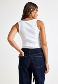 Hvid, ribbet tanktop med rund halsudskæring, parret med mørkeblå denimjeans, der har to baglommer og gule stikninger.