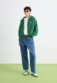 Green zip-up hoodie over a white t-shirt, paired with high-waisted blue jeans and white sneakers with green laces, standing on green flooring.