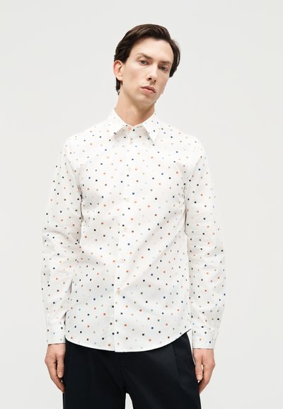 Άντρας που φοράει λευκό πουκάμισο με κουμπιά και πολύχρωμα μικρά τετράγωνα σχέδια και μαύρο παντελόνι, μπροστά σε απλό φωτεινό φόντο.