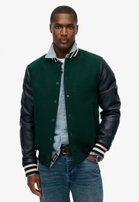 Varsityjack met een groene wollen body en marineblauwe leren mouwen, gestreepte geribbelde kraag, drukknoopsluiting en zijzakken, gedragen over denim.