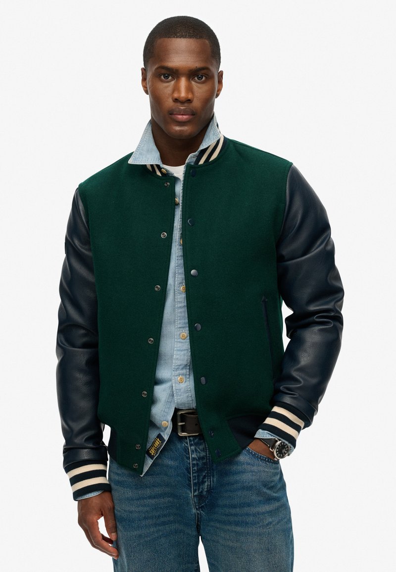 Varsityjack met een groene wollen body en marineblauwe leren mouwen, gestreepte geribbelde kraag, drukknoopsluiting en zijzakken, gedragen over denim.