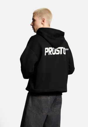 Młoda osoba z krótkimi blond włosami nosi czarną bluzę z kapturem z napisem „PROSTO” i symbolem nieskończoności na plecach, w połączeniu z ciemnymi dżinsami.