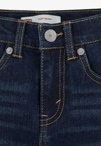 Tumši zili džinsi ar šauru krojumu, aprīkoti ar sudraba pogas aizdari, dzeltenām šuvju akcentiem un markas birku ar uzrakstu "510™ SKINNY".