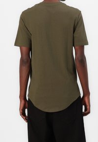 Camiseta de algodón en verde oliva, de manga corta, con dobladillo redondeado y cuello tipo barco. Sin patrones ni diseños visibles, silueta simple y entallada.