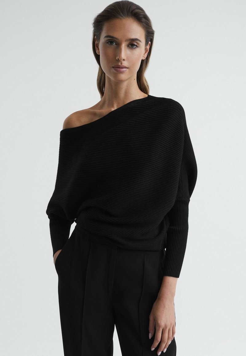 Zwarte geribbelde off-the-shoulder trui met lange mouwen, losse pasvorm en textielstof. Gecombineerd met zwarte high-waisted broeken.