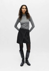 Femme aux longs cheveux bouclés portant un col roulé gris, une jupe portefeuille noire, des collants noirs et des bottes en cuir noires montantes, debout les mains derrière le dos.