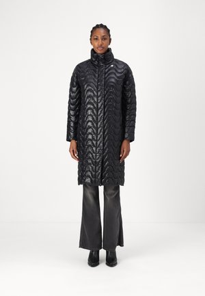 Black quilted long coat with a high collar, featuring wave-like stitching patterns. Worn over flared dark jeans and black ankle boots.