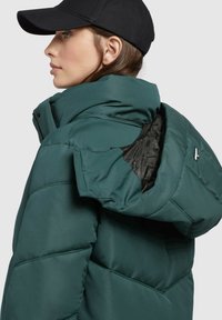 Jachetă puffer verde cu guler înalt, material texturat și căptușeală interioară neagră. Prezintă un închizător cu fermoar și o glugă.