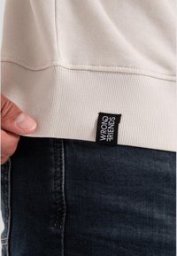 Light beige sweatshirt with ribbed hem, partially lifted to reveal a black label with white text. Paired with dark denim jeans.