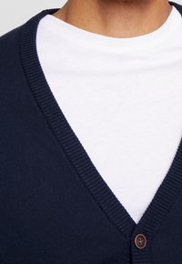 Námořnický modrý kardigan s výstřihem do V, ribovaným lemem a jedním hnědým knoflíkem na otevření, nošený přes bílý tričko.