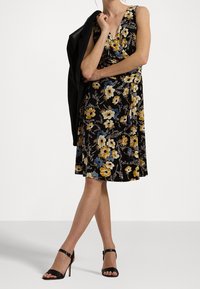 Mujer vestida con un vestido negro sin mangas con estampado floral y sandalias de tacón negras, sosteniendo una chaqueta negra sobre su hombro, de pie con las piernas cruzadas.