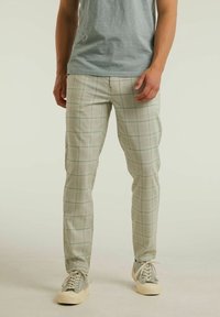 Man som bär ljusgrå rutiga byxor, ljusgrå t-shirt och grå sneakers med vita sulor, står framför en neutral bakgrund.