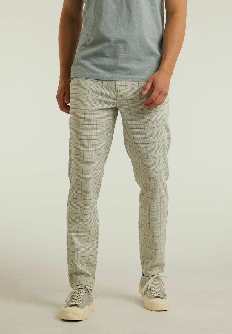 Man som bär ljusgrå rutiga byxor, ljusgrå t-shirt och grå sneakers med vita sulor, står framför en neutral bakgrund.