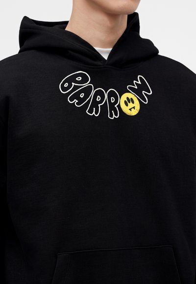 Personne portant un sweat à capuche noir avec le mot "BARROW" et un emoji visage triste jaune imprimé sur la poitrine près de l'encolure.