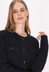 Cardigan a maglia cable di un blu navy scuro, caratterizzato da un collo rotondo, chiusure con bottoni e due tasche frontali con dettagli dorati. Texture morbida, vestibilità comoda.