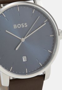 Reloj de hombre con esfera azul, índices plateados, indicador de fecha a las seis en punto y correa de cuero marrón. Caja de acero inoxidable.
