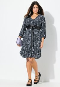 Schwarzes und weißes Kleid mit Blumenmuster und 3/4-Ärmeln, tailliert mit einem schwarzen Gürtel in der Taille, kombiniert mit einer lavendelfarbenen Clutch und Plateausandalen.