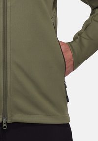 Olivgrüne Softshelljacke mit glatter Textur, vollem Reißverschluss und seitlichen Taschen. Schwarze Abschlüsse am Saum und um die Taschen.