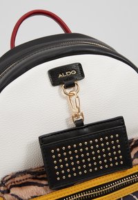 Primer plano de un bolso texturizado en blanco y negro con un asa roja, cremallera dorada, detalle de estampado de tigre y una etiqueta negra con studs adjunta debajo del logo de ALDO.