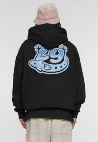 Sudadera negra con un gran gráfico azul en la parte trasera que muestra el número "29", un cohete y estrellas. El material parece suave y texturizado.