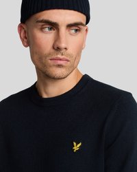 Jaunas vyras, vilkintis tamsiai mėlyną megztinį su geltonu paukščio logotipu ir megztiniu, žiūri šiek tiek į šoną prieš pilką foną.