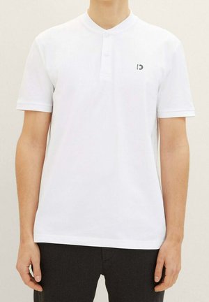Homme portant un polo blanc uni à manches courtes avec un petit logo noir sur la poitrine, associé à un pantalon sombre, devant un fond neutre.