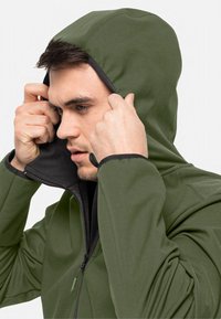 Veste à capuche verte en tissu lisse, avec un intérieur noir. Le mannequin ajuste la capuche avec une expression concentrée.