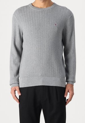 Mann trägt hellgrauen strukturierten Pullover mit kleinem rot-weiß-blauem Logo auf der Brust, kombiniert mit schwarzen Hosen, steht vor einfarbigem Hintergrund.