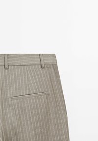 Beige streepjesbroek met riemlussen en een enkele achterzak, tentoongesteld tegen een witte achtergrond.