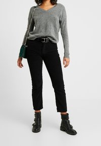 Pull gris à col en V, pantalon en velours côtelé noir taille haute et longueur cheville, bottines noires avec boucles, sac à main vert avec chaîne.