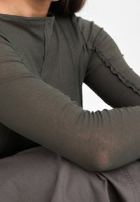Dark green long-sleeve top with ribbed texture, featuring a round neck and slight ruffle detail along the sleeve seams.