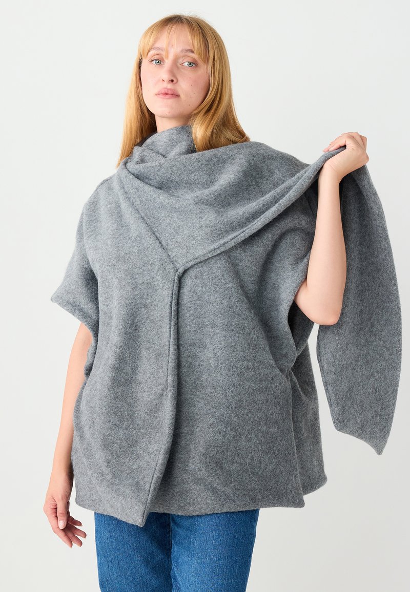 Poncho en laine grise aux manches oversized, avec un col drapé et une finition texturée. Porté légèrement ouvert à l'avant, maintenu d'une main.