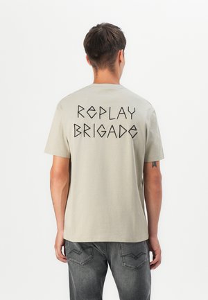 Šviesiai rudos spalvos trumpomis rankovėmis marškinėliai su apvalia iškirpte, ant nugaros esantis juodas geometrinis tekstas "REPLAY BRIGADE". Pagaminti iš minkštos medvilnės medžiagos.
