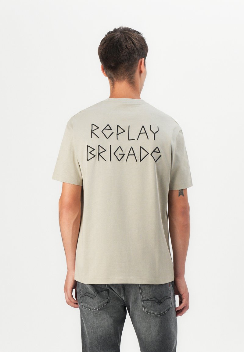 Krátkozrnný béžový tričko s kulatým výstřihem, na zadní straně s černým geometrickým textem "REPLAY BRIGADE". Vyrobeno z měkkého bavlněného materiálu.