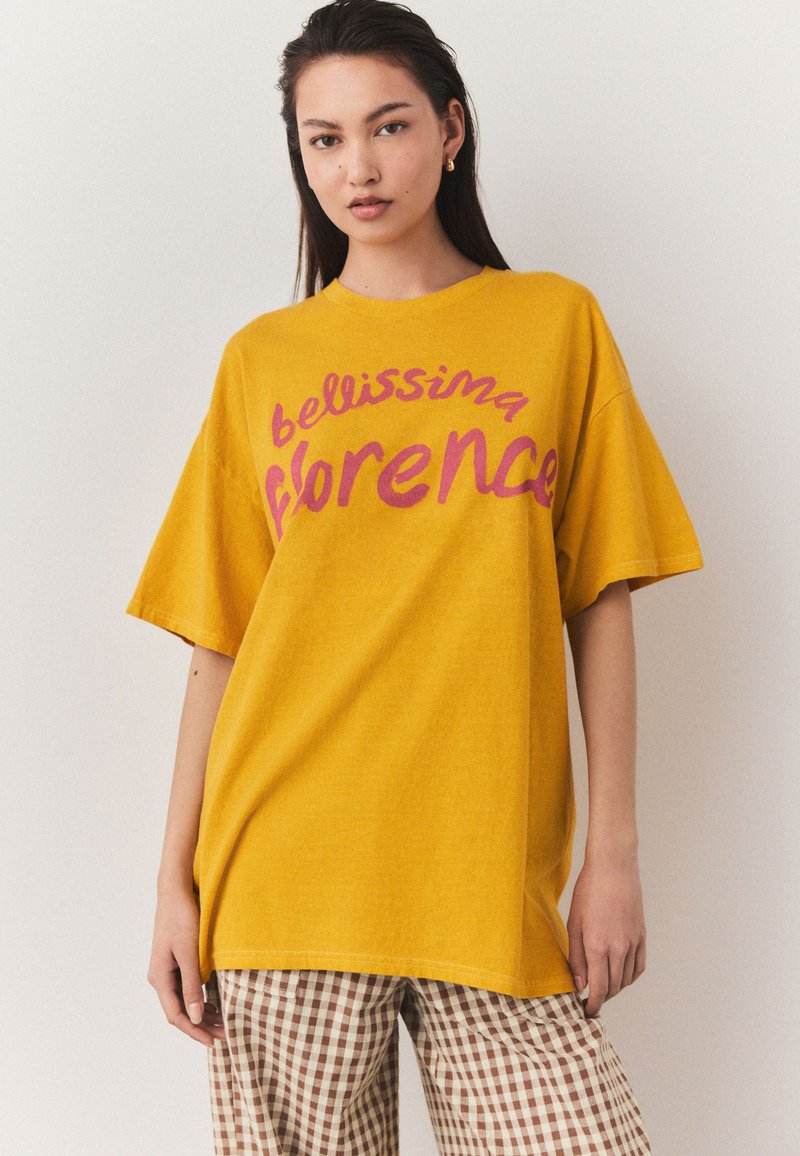 Maglietta gialla oversize con la scritta "bellissima Florence" in testo rosa. Presenta maniche corte e una vestibilità rilassata, abbinata a pantaloni a quadri.