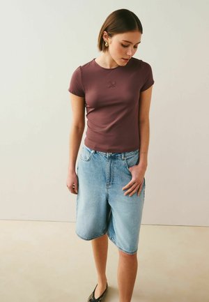 Fiatal nő barna, testhez simuló, rövid ujjú felsőt és világoskék farmer bermuda sortot visel, egyik kezét a zsebében tartja.