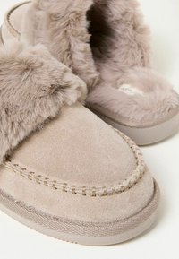 Fuzzy beige slippers with a soft suede exterior, featuring plush fur around the collar and decorative stitching on the upper.