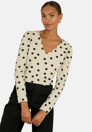 Vrouw draagt een beige top met lange mouwen, zwarte polka dots en een gekartelde halslijn, gecombineerd met zwarte, hooggetailleerde broek, poserend tegen een witte achtergrond.