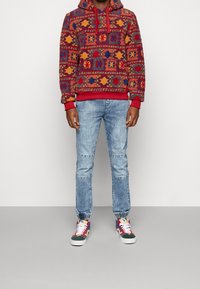 Červená a vzorovaná mikina s kapucí a stahovací šňůrkou, v kombinaci se světle modrými džíny s nášivkami na kolenou. Pestré tenisky s bílými tkaničkami.