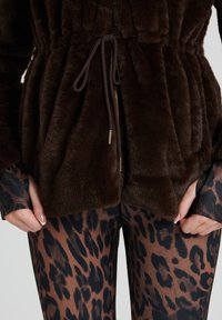 Brun fuskpälsjacka med elastisk midja, ficka med dragkedja och dragsko. Matchad med bruna leggings i leopardmönster med en slät textur.