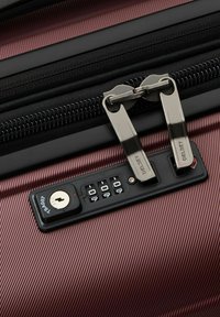 Valise marron avec texture côtelée, équipée d'une fermeture éclair noire et de tirettes doubles en argent. Comprend un cadenas à combinaison et un trou de clé.