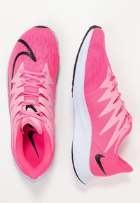 Zapatillas deportivas rosas con parte superior de malla texturizada, que presentan un logo de swoosh negro, acentos en zigzag y una entresuela blanca con suela de goma.
