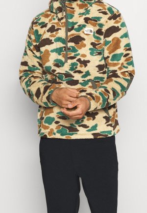 Person trägt einen Fleece-Pullover mit Camouflage-Muster in Grün-, Braun- und Beigetönen und schwarze Hose, Hände vor dem Körper verschränkt.
