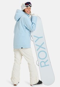 Helles blaues Schneejacke mit Kapuze, kombiniert mit weißen Schneehosen und schwarzen Snowboardstiefeln. Hält ein weißes Snowboard mit dem "ROXY"-Logo.