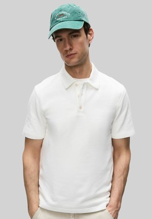 Ung mand iført en hvid kortærmet poloshirt og grøn kasket med teksten "Slow Sundays Since 1974" mod en ensfarvet grå baggrund.