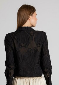Zwarte doorschijnende blouse met geborduurd bloempatroon, lange mouwen en een kraag. De achterkant toont gestructureerde details en een getailleerd silhouet.