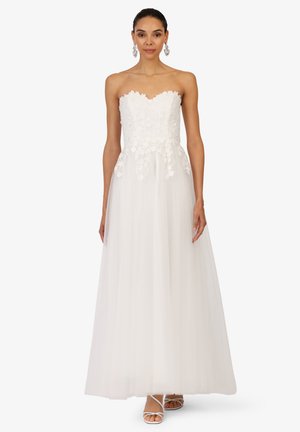 Weißes trägerloses Kleid mit einem floralen Spitzenoberteil, Tüllrock und einem herzförmigen Ausschnitt. Das Model trägt silberne Ohrringe und weiße Sandalen mit Riemchen.